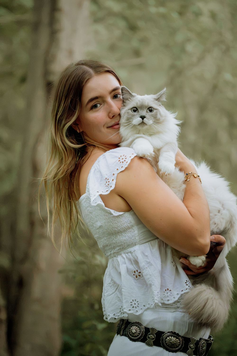Young woman in white eyelet dress holds a fluffy Ragdoll cat outdoors in a lush green forest setting.