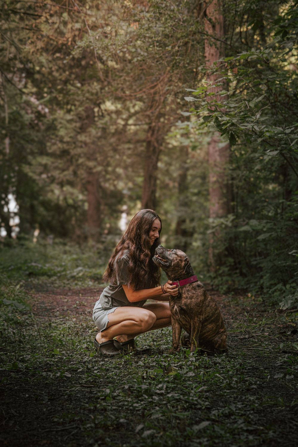 A young woman crouches on a forest trail, smiling while petting her brown dog wearing a red collar.