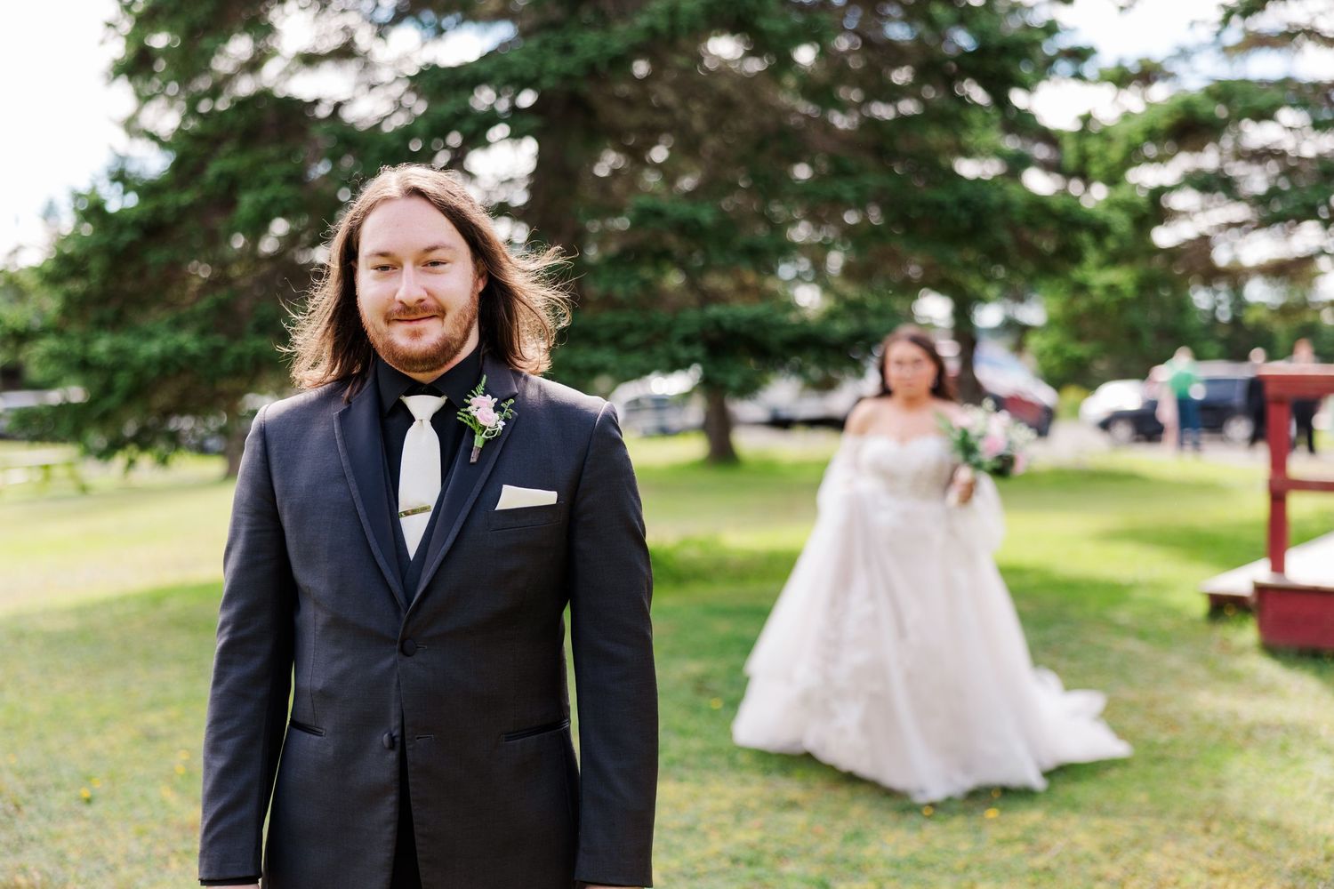 Groom in black suit smiles outdoors as bride in white gown approaches during wedding first look ceremony.