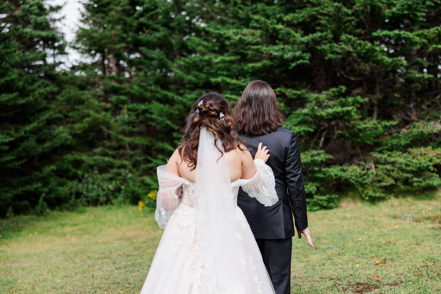Bride in lace gown and groom in black suit walk arm in arm toward evergreen trees at outdoor wedding.