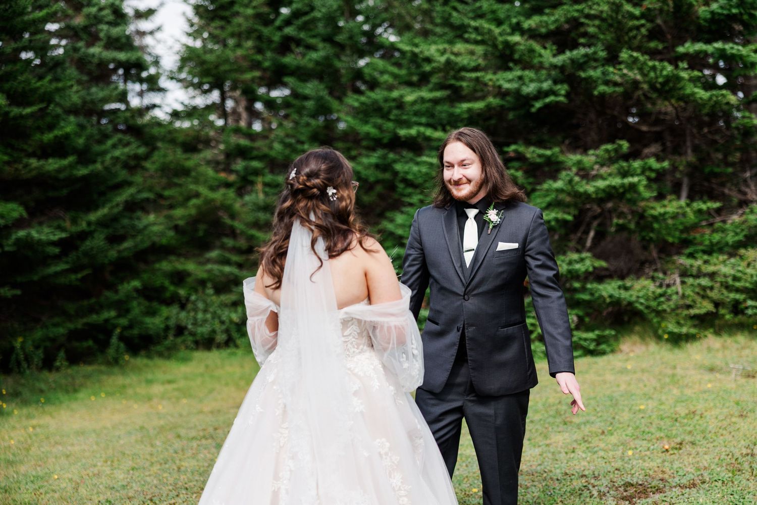 A bride in a white gown and groom in a black suit share a joyful first look moment outdoors among tall green pine trees.