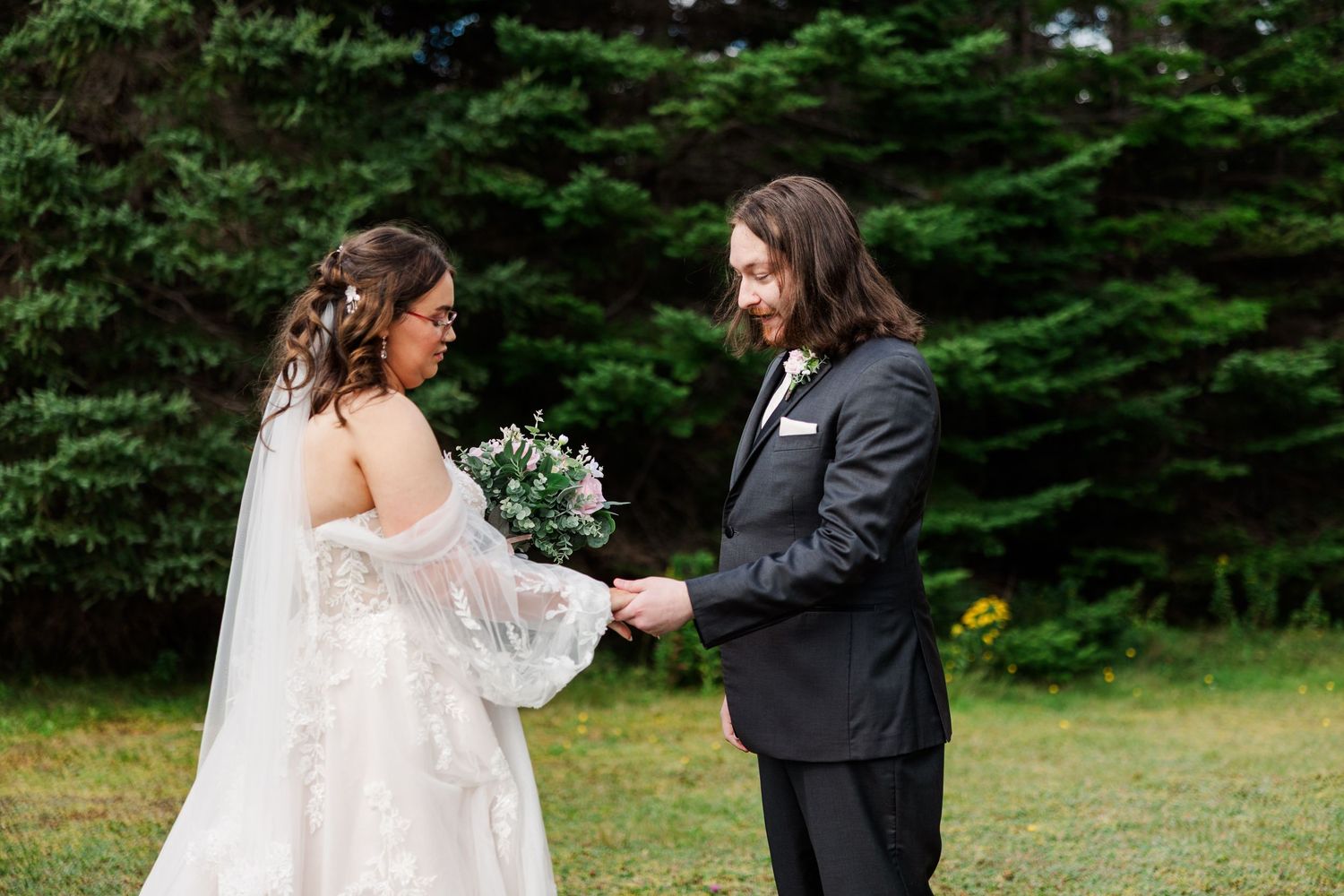 Bride in white gown holding bouquet exchanges vows with groom in black suit outdoors near evergreen trees.