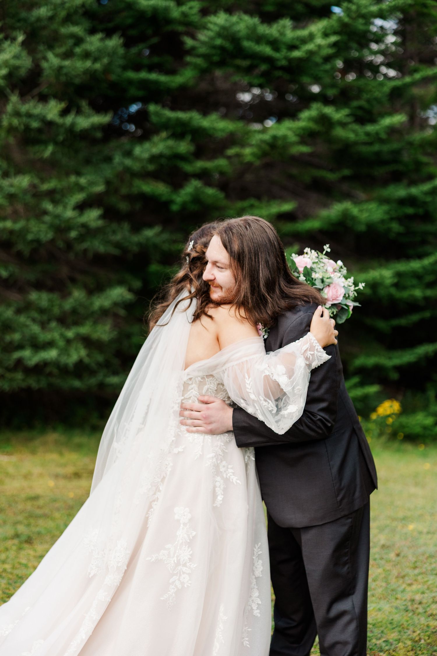 Bride and groom embracing outdoors, she holds a pink bouquet, wearing lace gown with veil, he wears black suit, evergreen trees behind.