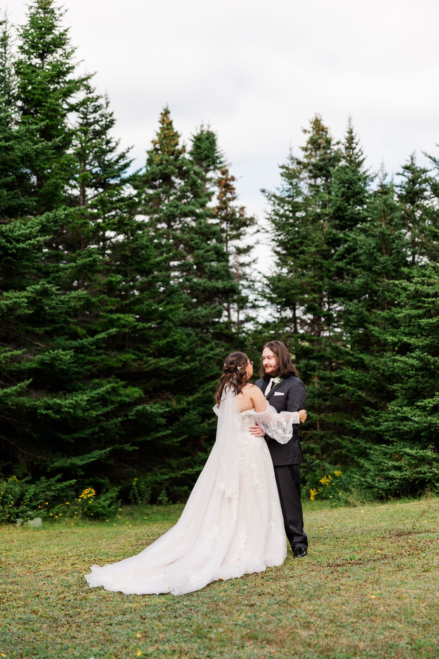 A bride and groom share a romantic embrace on a green lawn surrounded by tall evergreen trees on their wedding day.