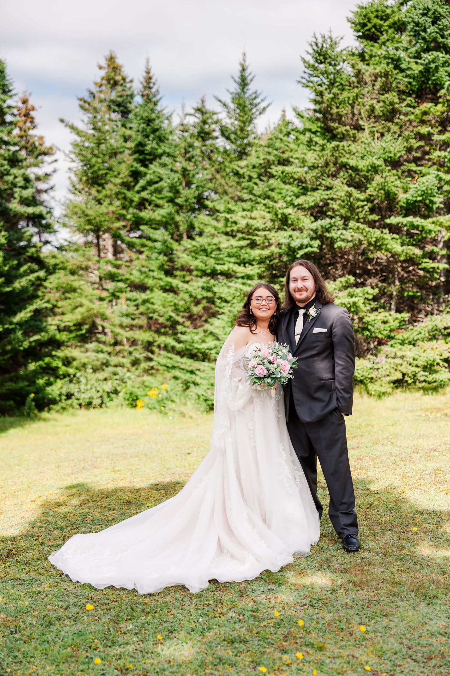 Bride in white gown holding pink bouquet stands with groom in black suit outdoors near evergreen trees on wedding day.