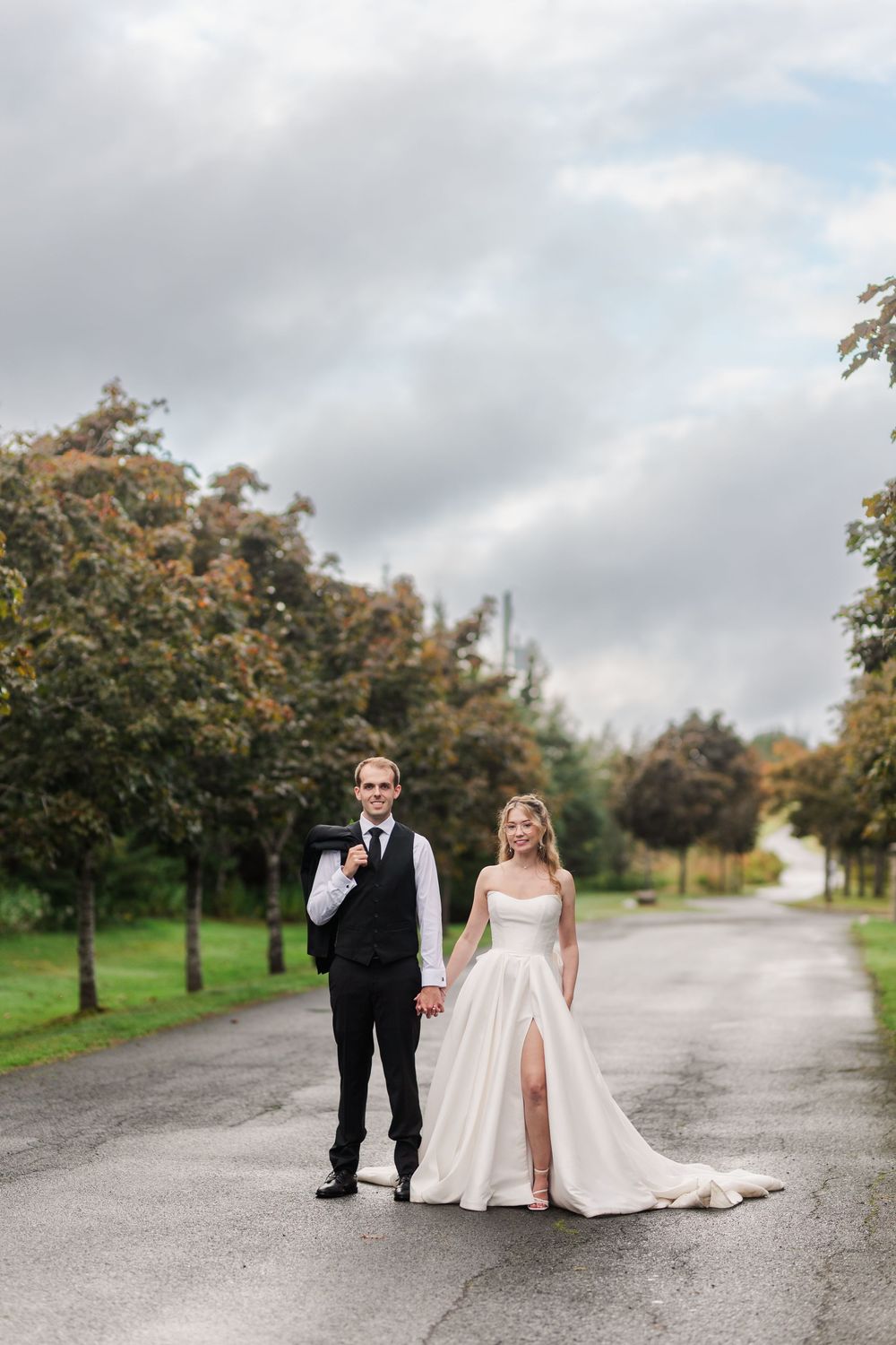 Bride and groom holding hands on a tree-lined road, bride in a white slit gown, groom in a black vest suit.