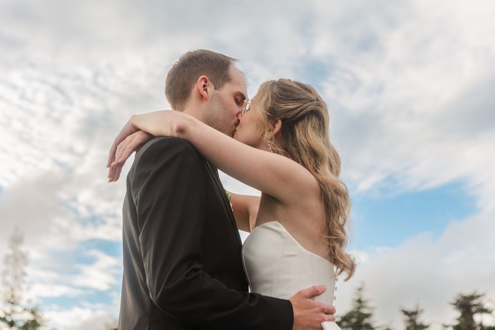 A bride and groom share a romantic kiss outdoors under a cloudy blue sky on their wedding day.