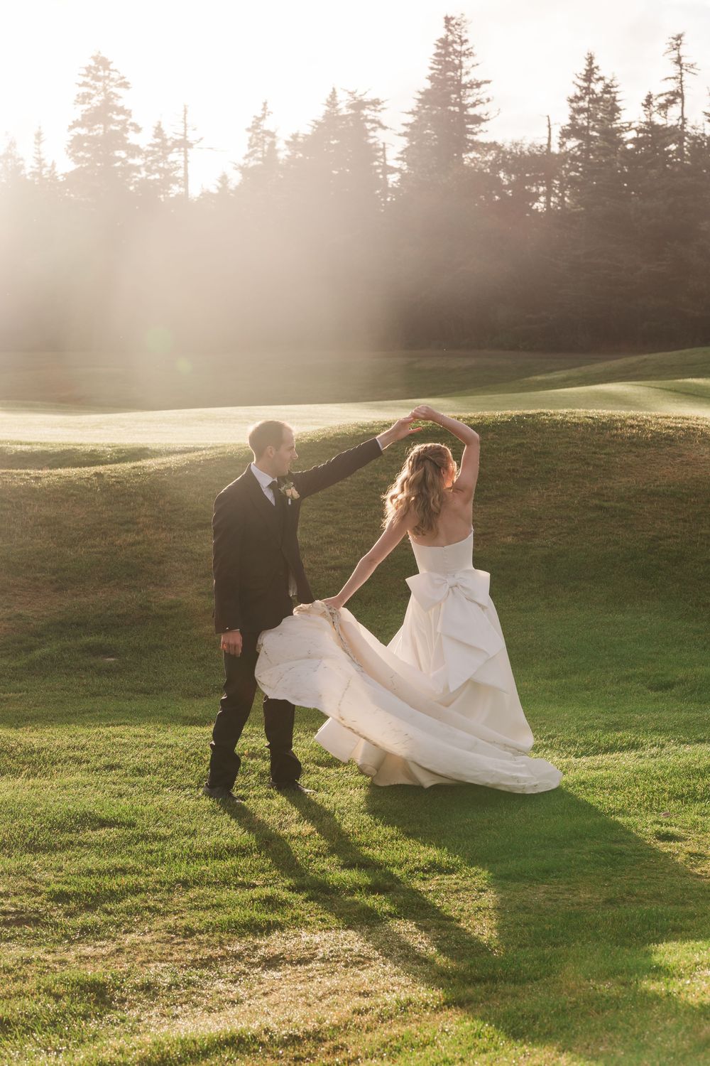 Bride and groom dancing outdoors on lush green golf course at golden hour, surrounded by pine trees.