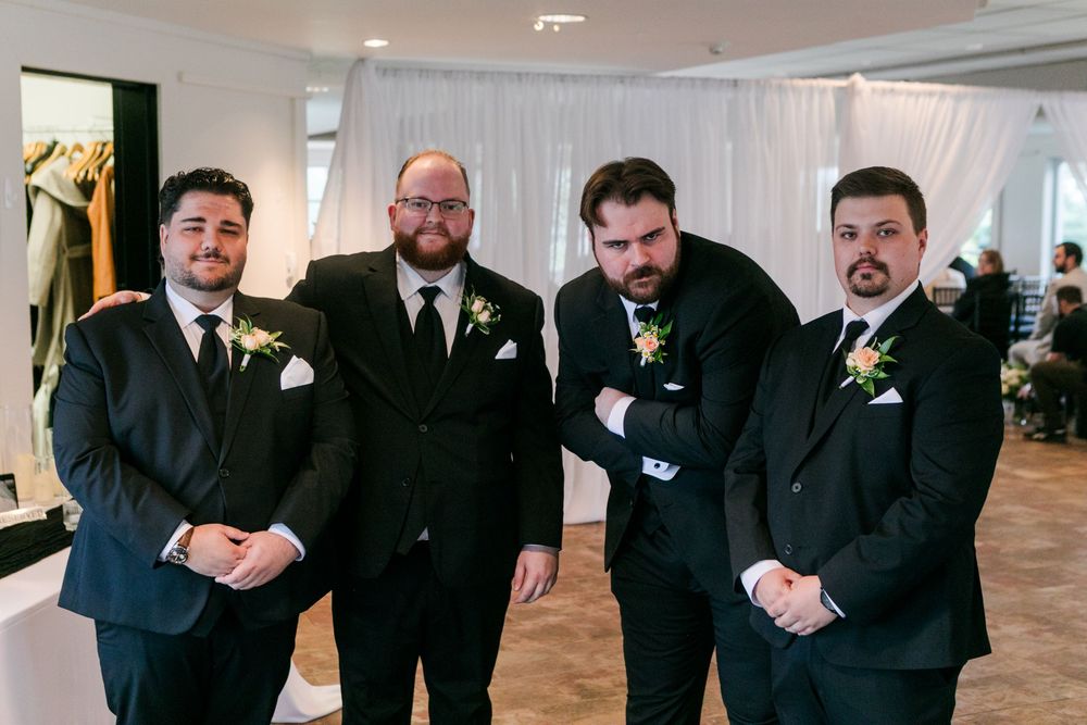 Four groomsmen in black suits with boutonnieres posing together at a wedding venue.