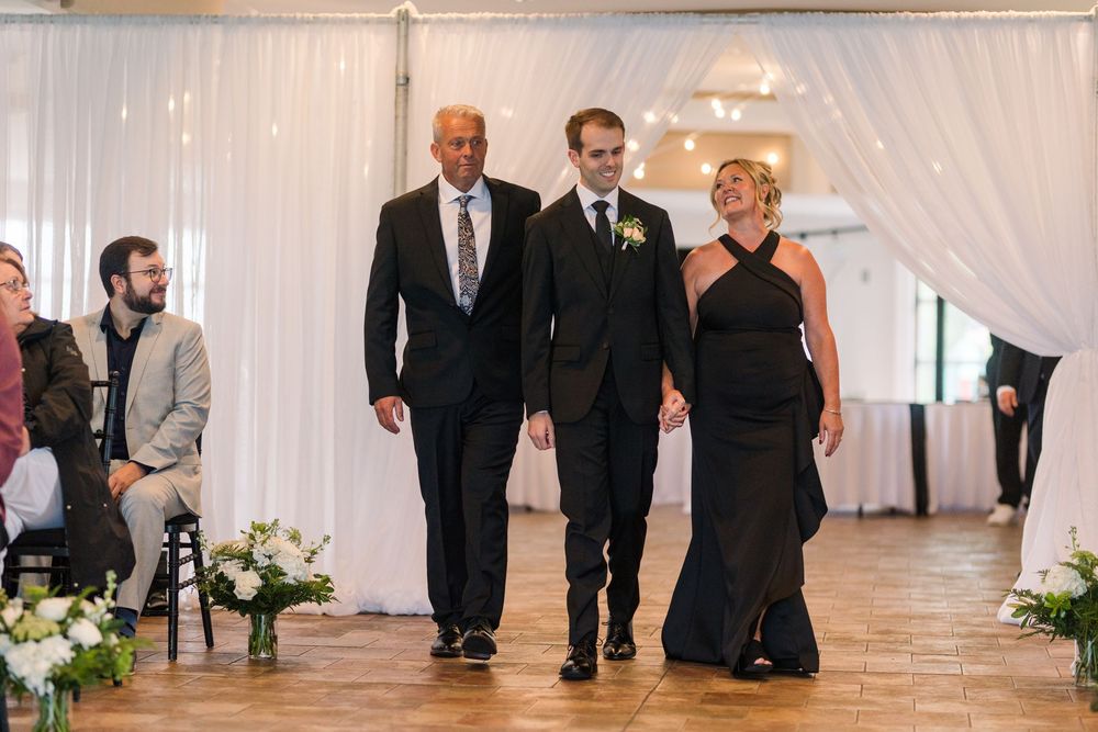 Groom escorted down the aisle by his parents at an elegant white-draped wedding ceremony.