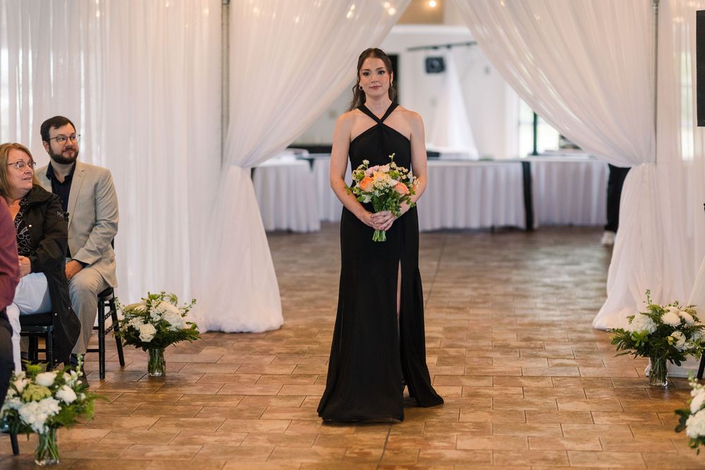 Bridesmaid in black halter gown walks down aisle holding colorful bouquet at elegant indoor wedding ceremony.