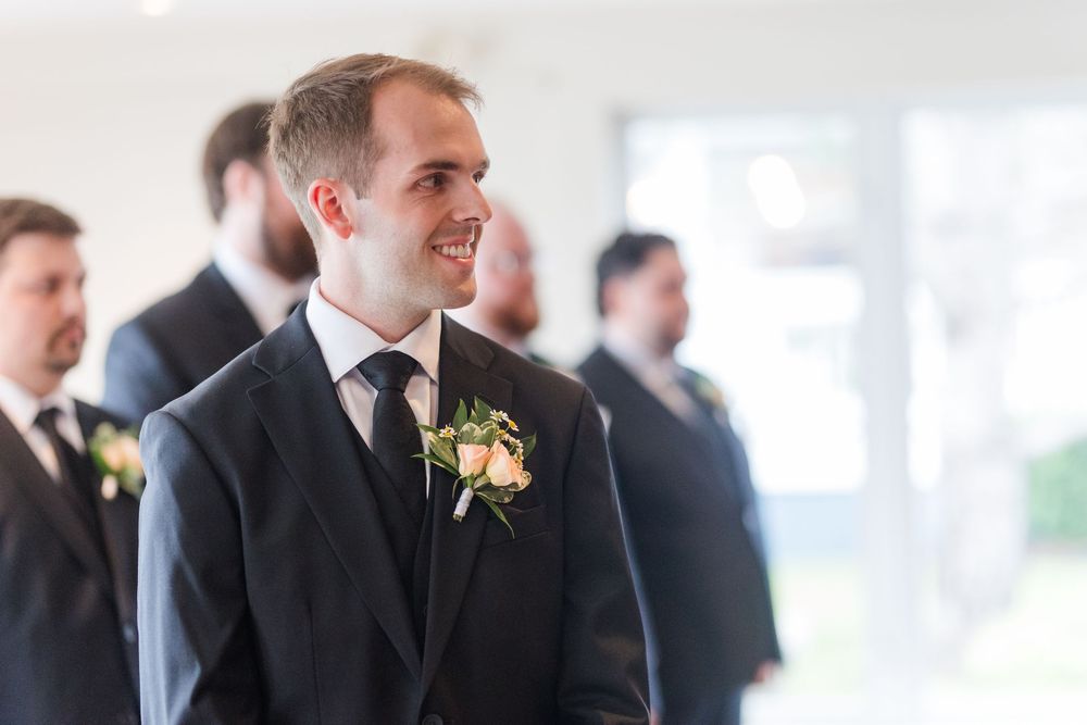 Smiling groom in dark suit with floral boutonniere stands at altar, groomsmen visible in background.