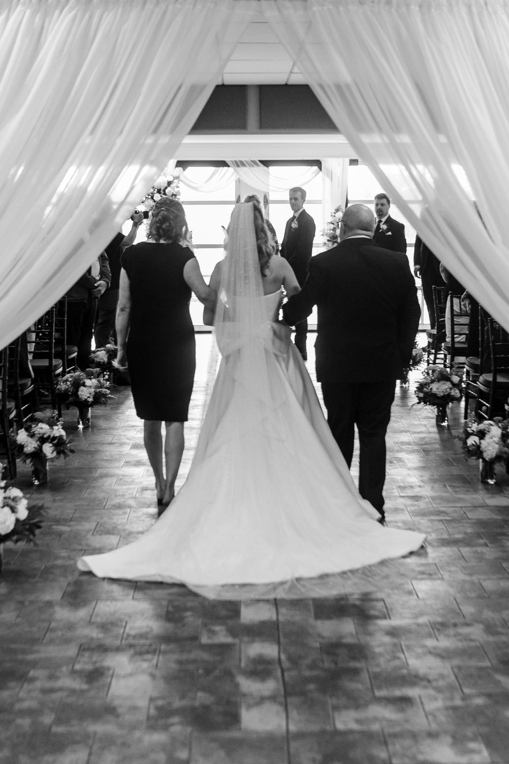 Black and white photo of a bride walking down the aisle escorted by two people at an elegant indoor wedding ceremony.