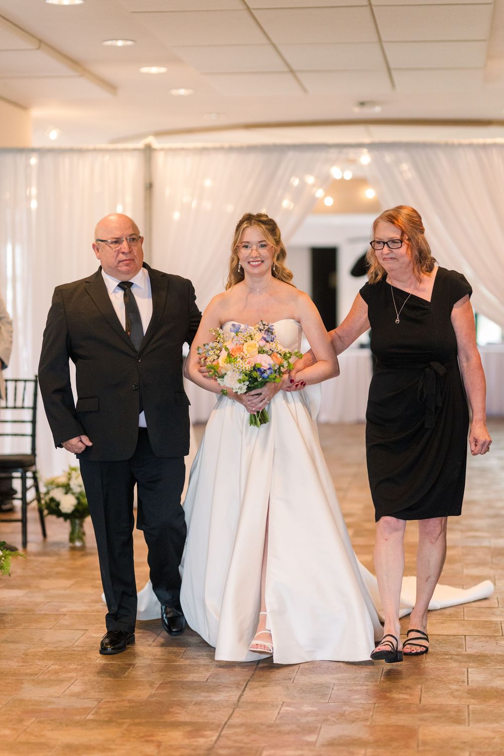 Bride in white gown walks down aisle with parents, holding colorful bouquet at elegant wedding venue.