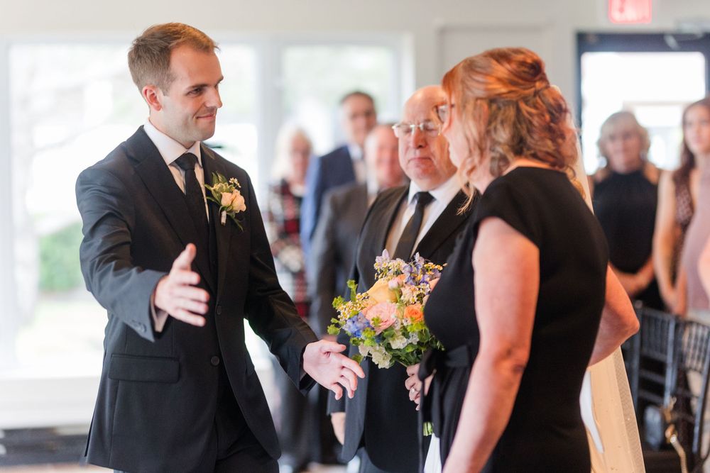 Groom in dark suit with boutonniere greeting guests holding flowers at an indoor wedding ceremony.