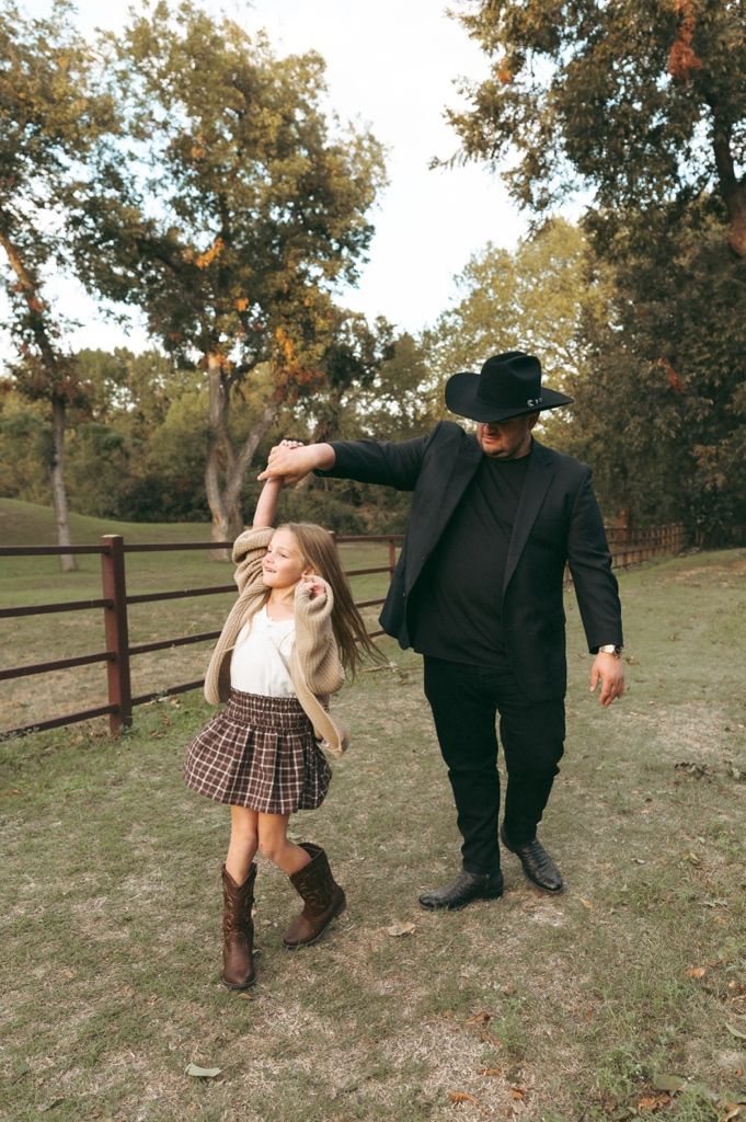 Dad twirling his little girl in a plaid skirt and boots on a grassy path by a wooden fence taken by Mary Hickman Photography.
