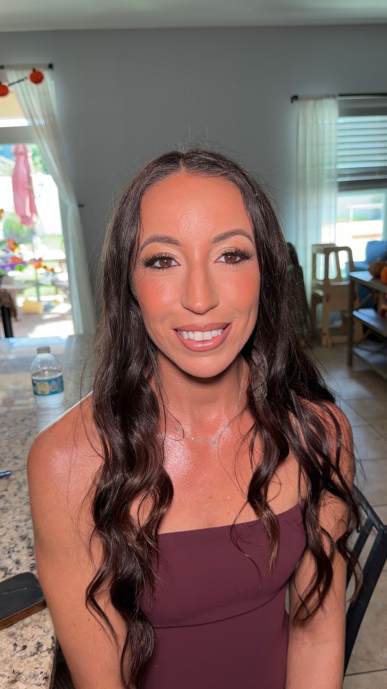 Smiling woman with long wavy brown hair and polished makeup wearing a burgundy off-shoulder dress, seated in a bright kitchen.