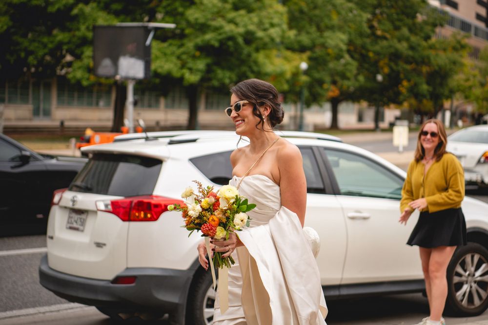Bride in an off-the-shoulder white gown and sunglasses holds a colorful bouquet beside a white SUV while a woman in yellow walks by.