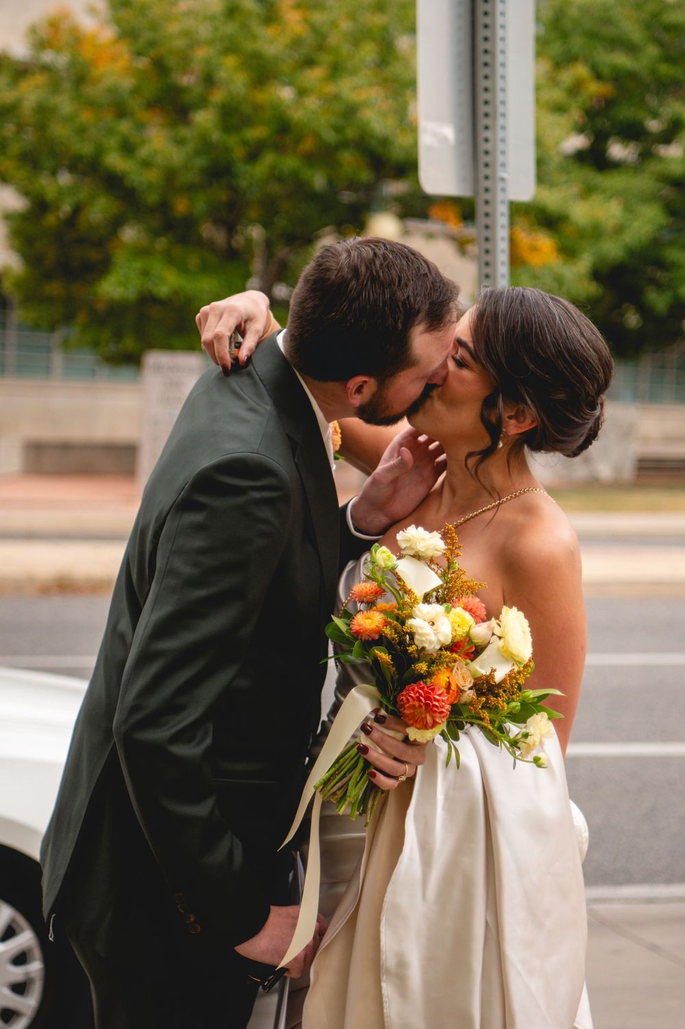 Bride in a satin gown holding an orange and white floral bouquet kisses her groom in a dark suit on a city street.