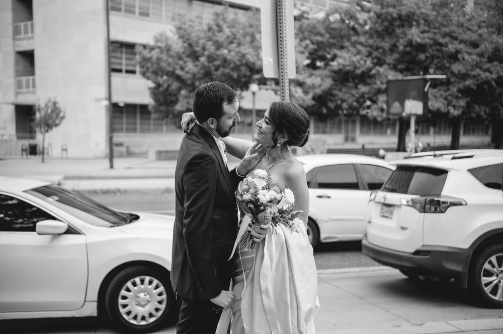Black and white photo of a bride holding a bouquet being embraced by a groom in a suit on a city street beside parked cars.