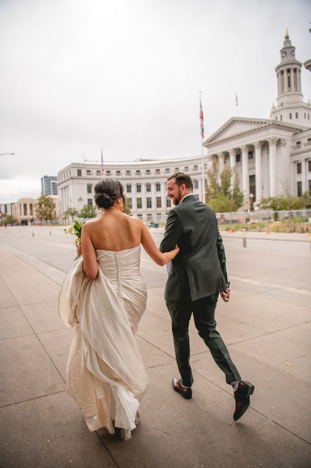 Bride in a strapless wedding gown and groom in a green suit walk hand in hand past a grand neoclassical government building.