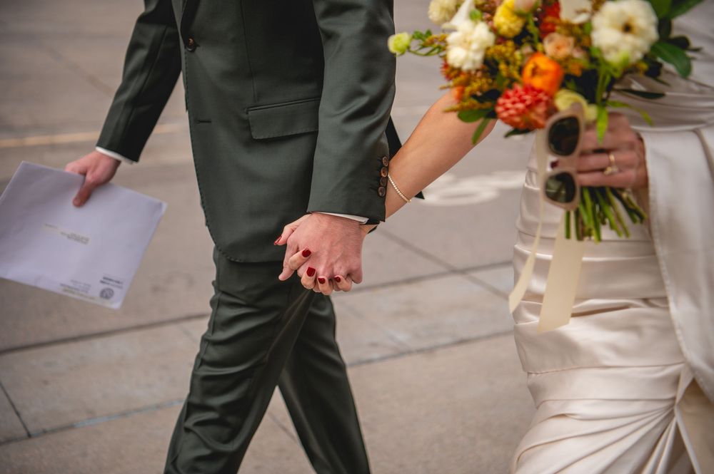 Bride in a white gown and groom in a dark suit hold hands while walking, with a colorful floral bouquet and paper in hand.