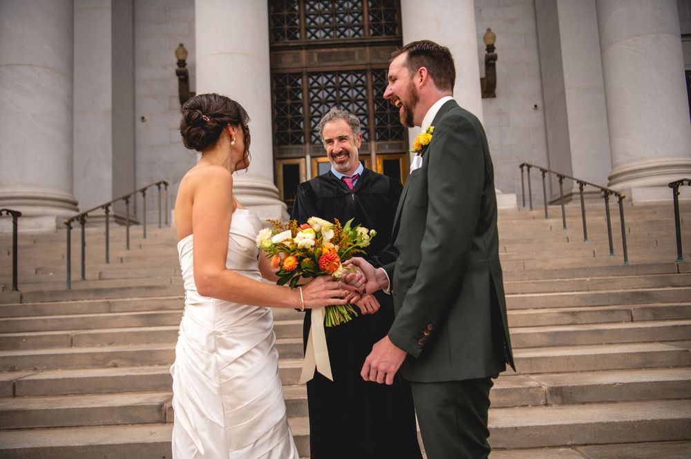 Bride in a white strapless gown holds an orange bouquet and joins hands with the groom as an officiant marries them on stone steps.