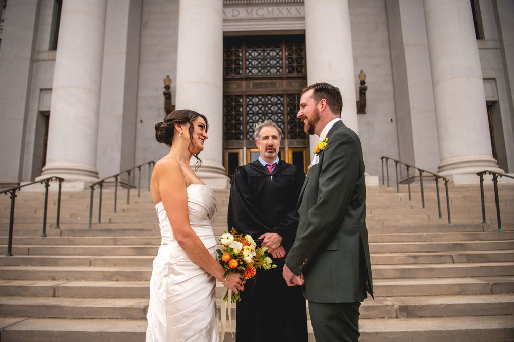 Bride in white gown holding an orange bouquet exchanges vows with a groom in a green suit as an officiant stands on courthouse steps.