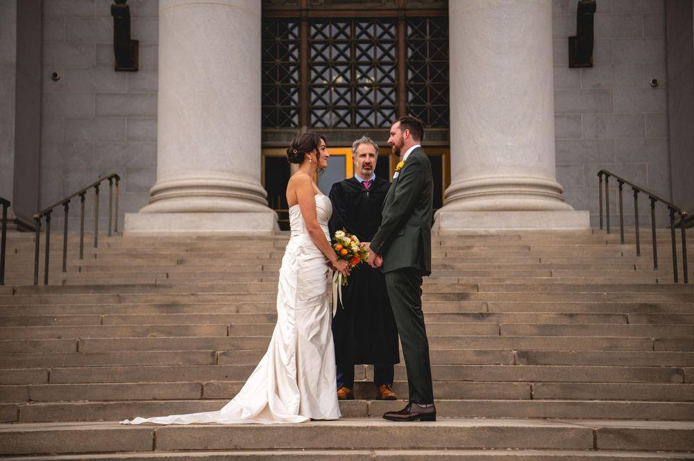 Bride in white gown and groom in dark suit hold a bouquet during an outdoor wedding ceremony on stone steps with an officiant and columns.