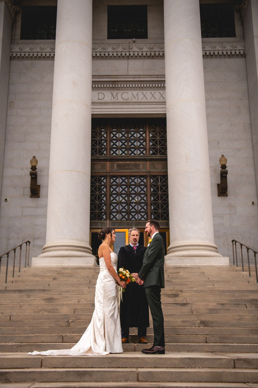 Bride in a white gown and groom in a green suit exchange vows with an officiant on the steps of a grand columned building.