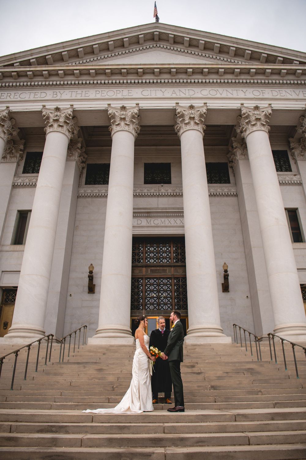 Bride and groom stand with an officiant on the steps of a grand neoclassical courthouse with tall stone columns during a wedding ceremony.