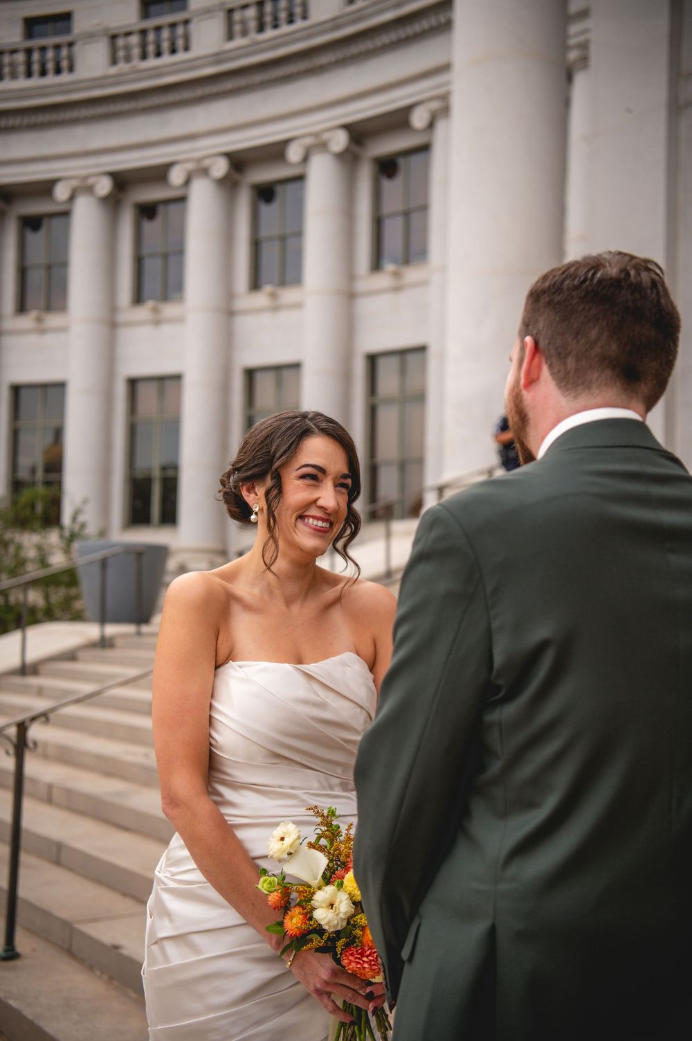 A smiling bride in a strapless white dress holds a colorful bouquet while facing a groom in a green suit on stone steps.