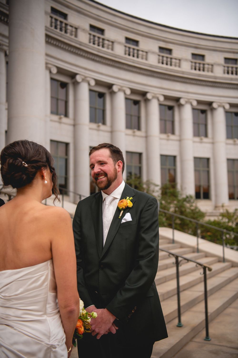 Smiling groom in a dark suit with an orange boutonniere faces his bride during an outdoor wedding ceremony before a grand columned building.