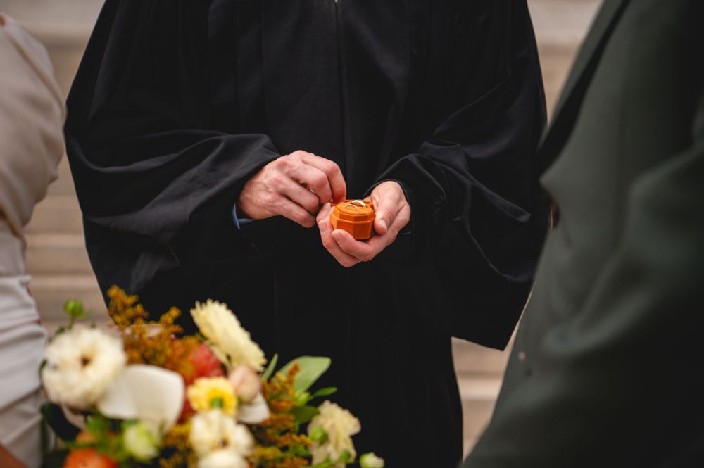 Officiant in a black robe holds a small orange rose in folded hands during a wedding ceremony, with a flower bouquet in the foreground.