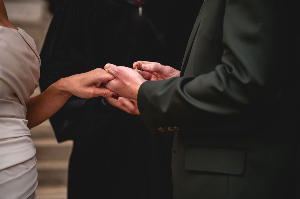 Couple joining hands during a wedding ceremony, with an officiant in dark robes standing behind them.
