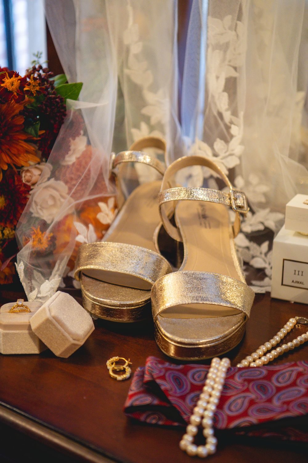 Gold platform wedding sandals beside pearl necklaces, gold rings, a paisley pocket square, ring boxes and autumn flowers on a wooden table.