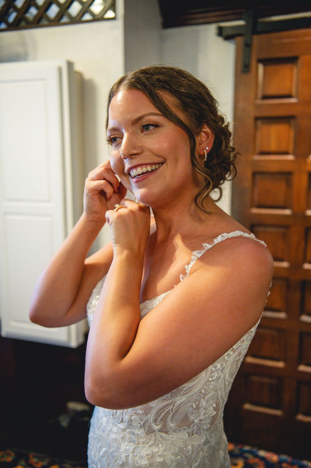 Smiling bride in a lace wedding gown adjusts her earring while getting ready in a room with wooden paneled doors.