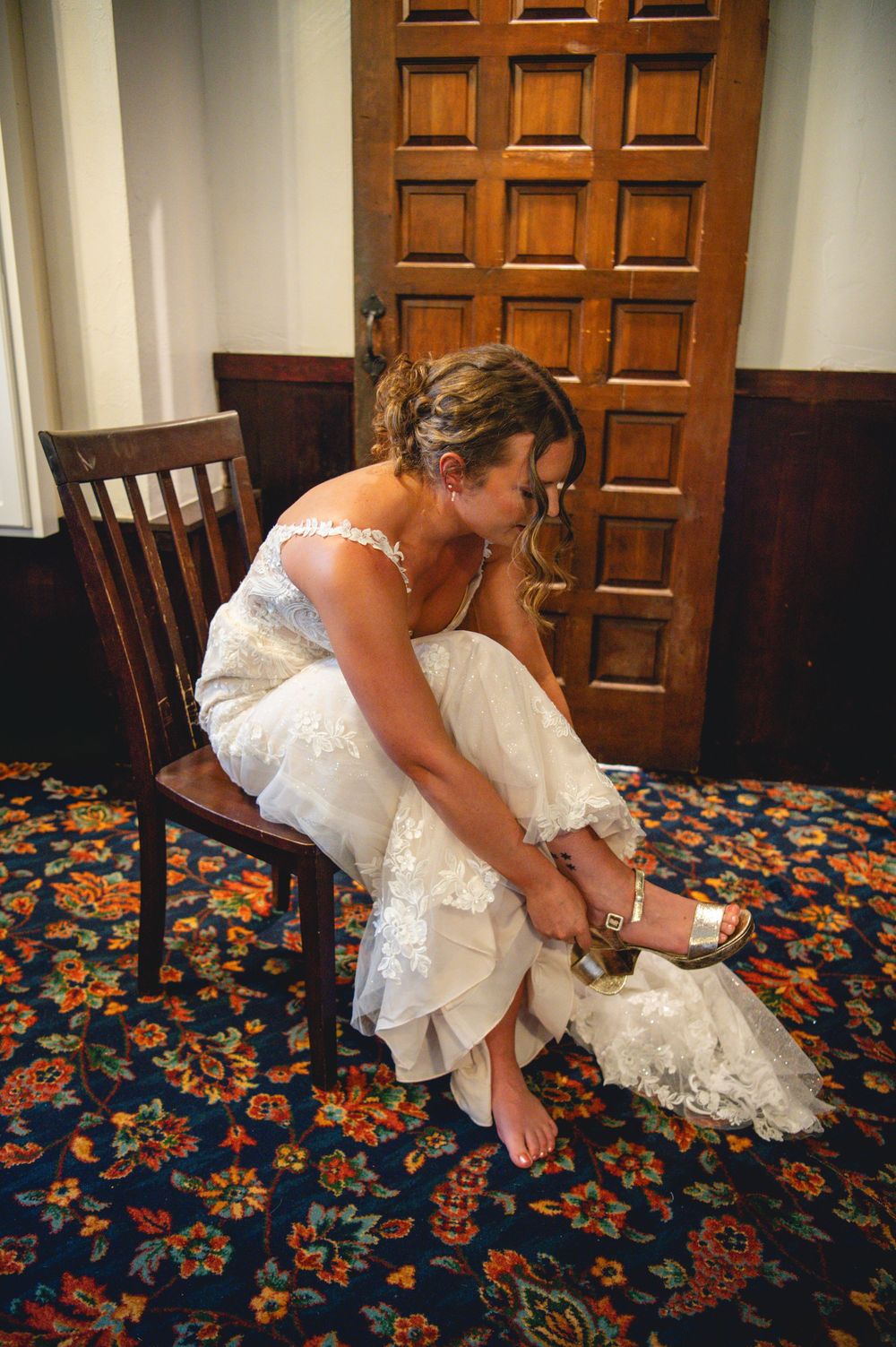 A bride in a lace wedding dress sits on a wooden chair, adjusting her gold sandal on a colorful patterned carpet before the ceremony.