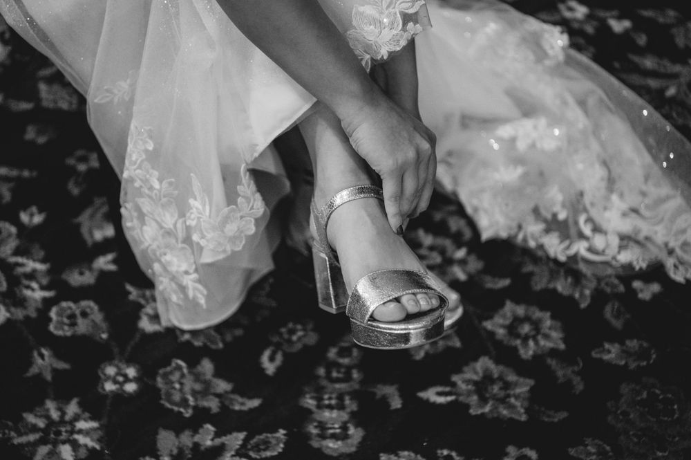 Bride in a lace wedding gown adjusts the strap of her silver platform sandal while seated on a patterned floral rug.