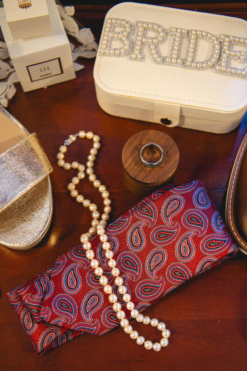 Bridal flat lay with a pearl-beaded BRIDE jewelry case, pearl necklace, silver ring in wooden box, paisley pocket square, and gold sandal.