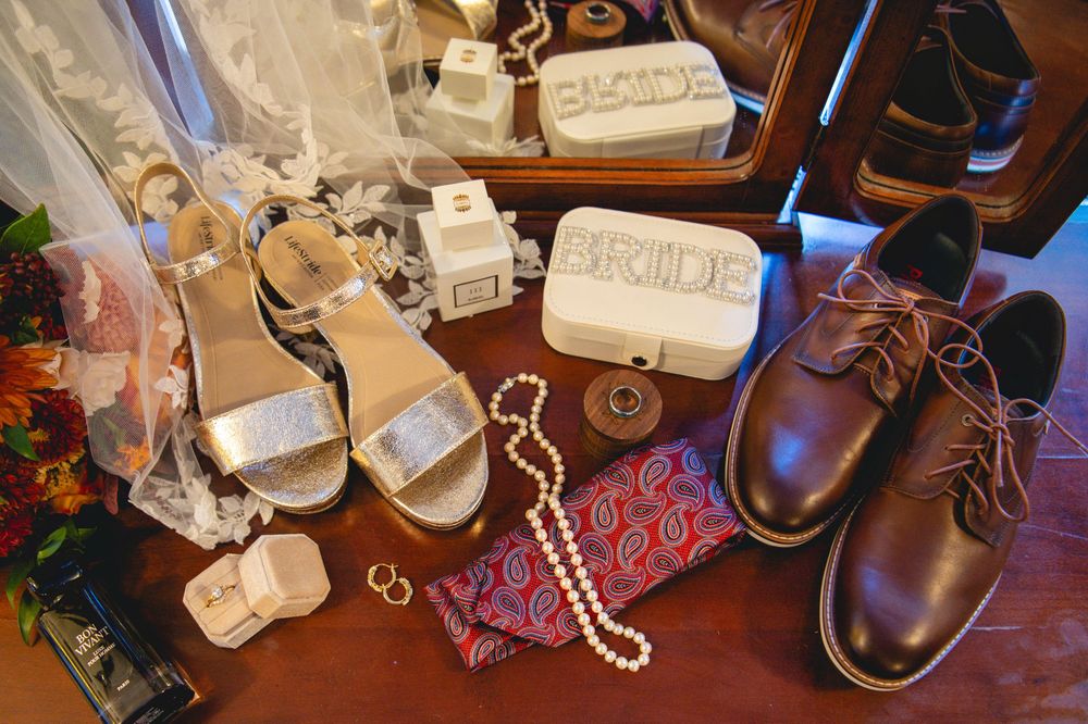 Wedding day flat lay with silver bridal sandals, pearl necklace, bride perfume bottle, brown groom shoes, red paisley pocket square and lace