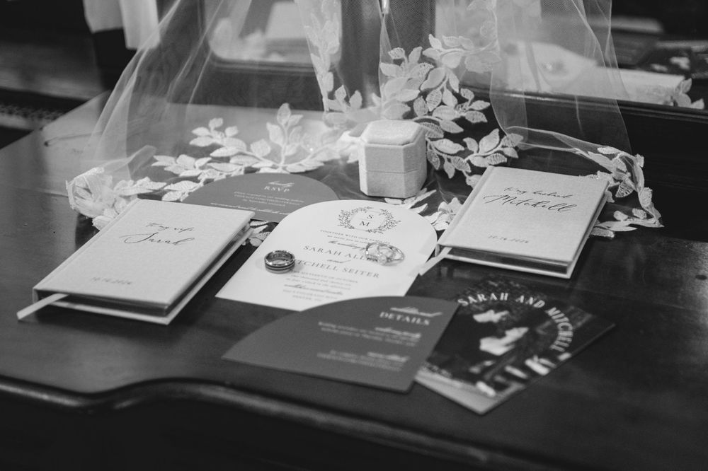 Black and white photo of wedding stationery, vow books, and a ring box laid on a table beneath a lace veil with floral details.