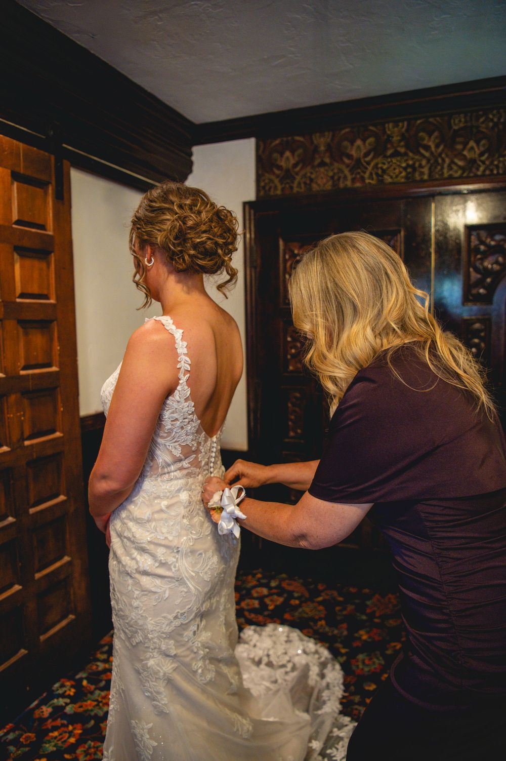 A bride in a lace wedding gown with an updo is helped with her dress by a woman in a plum top inside a wood-paneled room.