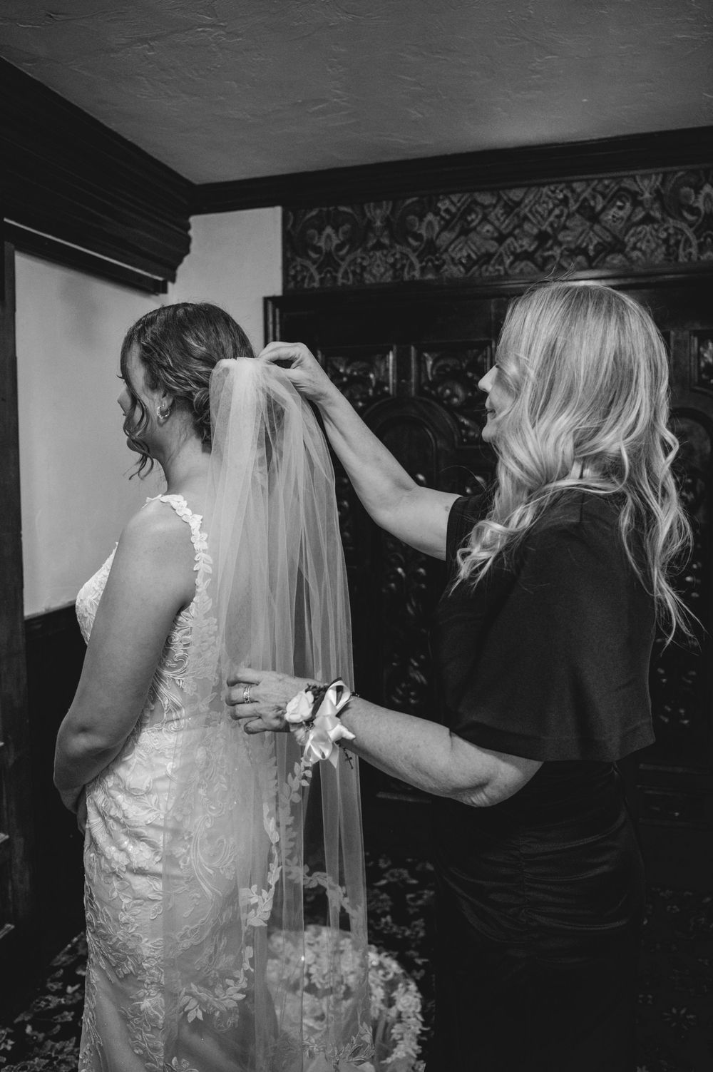A woman in a black dress carefully adjusts the lace veil of a bride in a white lace gown in a black and white wedding photo.