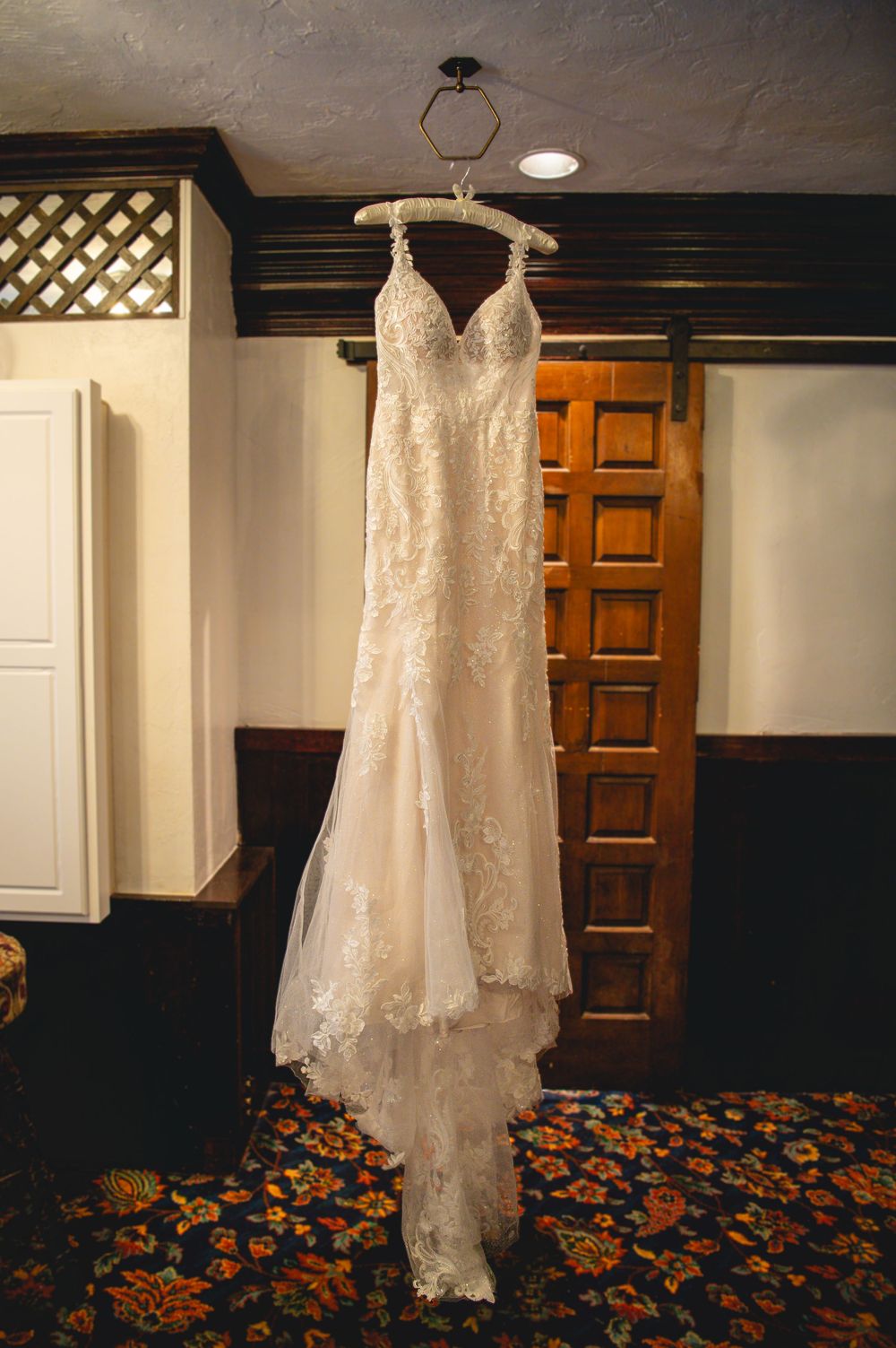 Lace wedding gown with illustrated straps and a flowing tulle skirt hangs from a geometric hanger beside a wooden door.