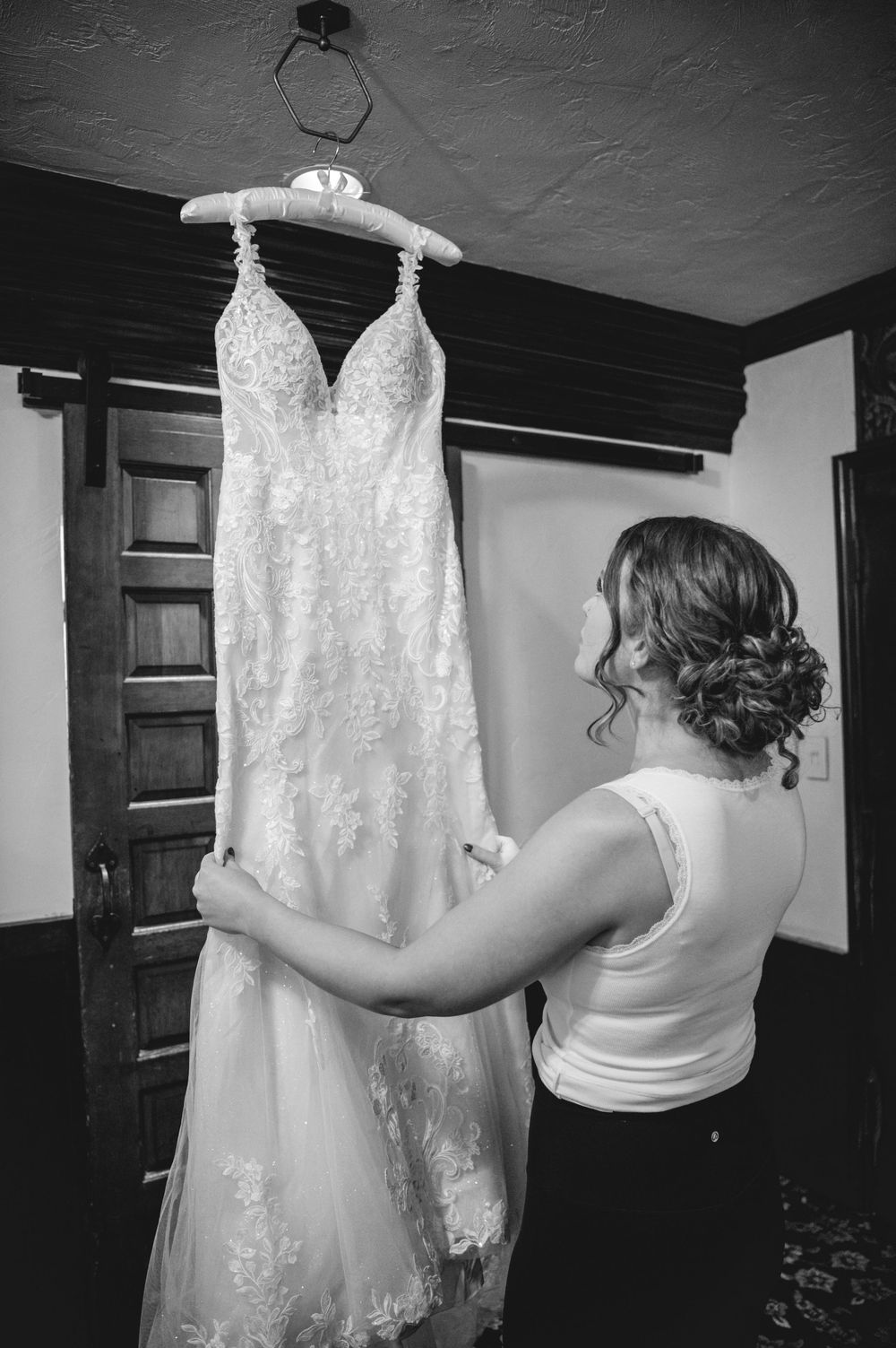 Bride in a white tank top gazes at a lace wedding gown hanging from a wooden door in a black and white photo.