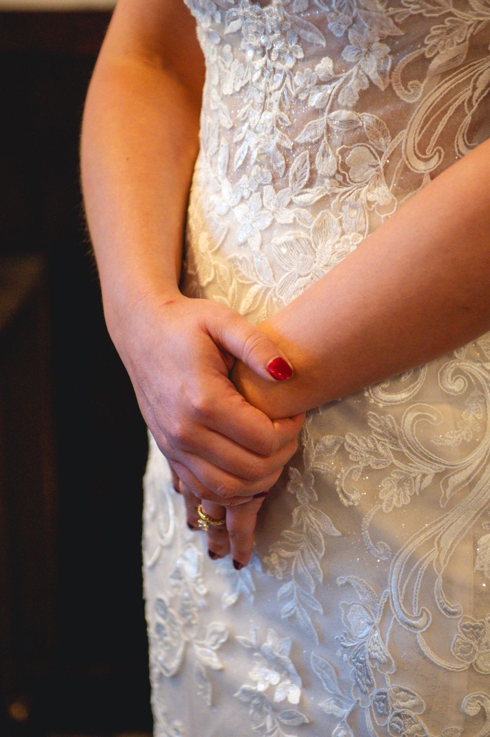 Close-up of a bride's clasped hands with red nail polish and a gold ring, resting on her lace wedding dress.