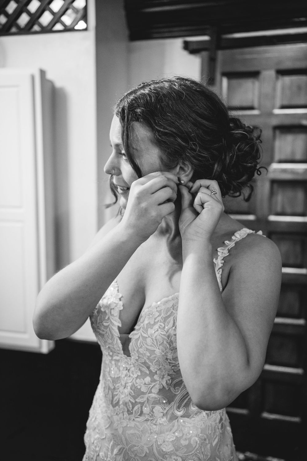 Black and white photo of a bride in a lace wedding gown with an updo, putting on an earring while getting ready.