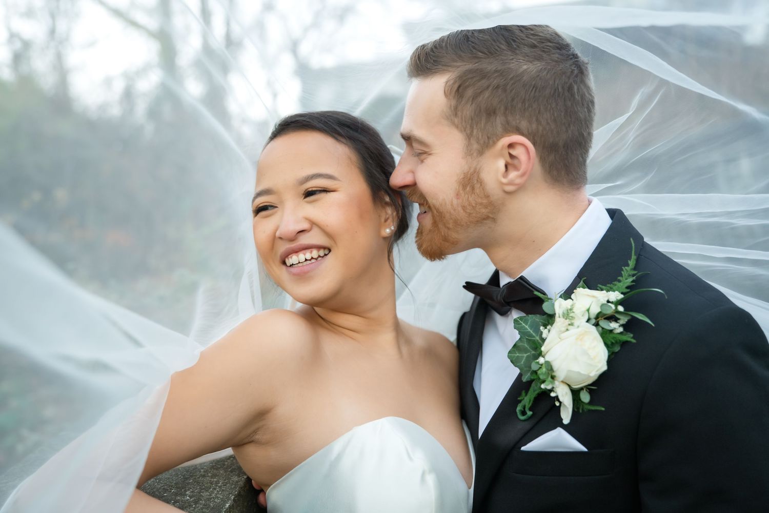 Bride in a white gown and groom in a gray blazer embrace in a grassy field overlooking the water at sunset.