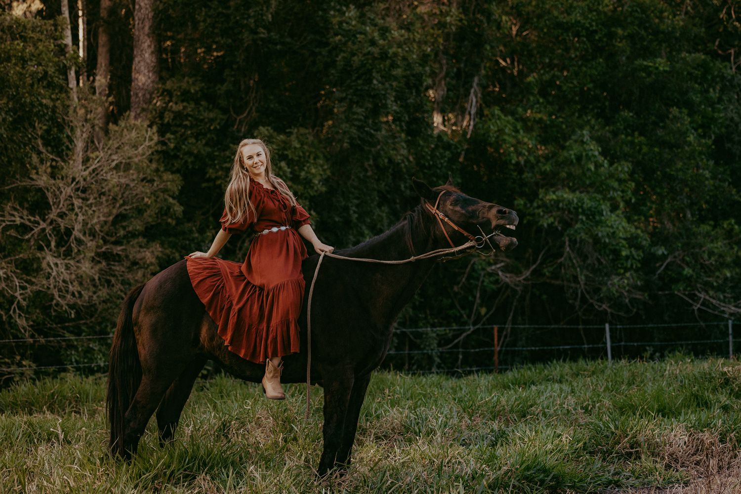 A woman in a flowing red dress sits gracefully atop a dark horse in a green countryside setting.